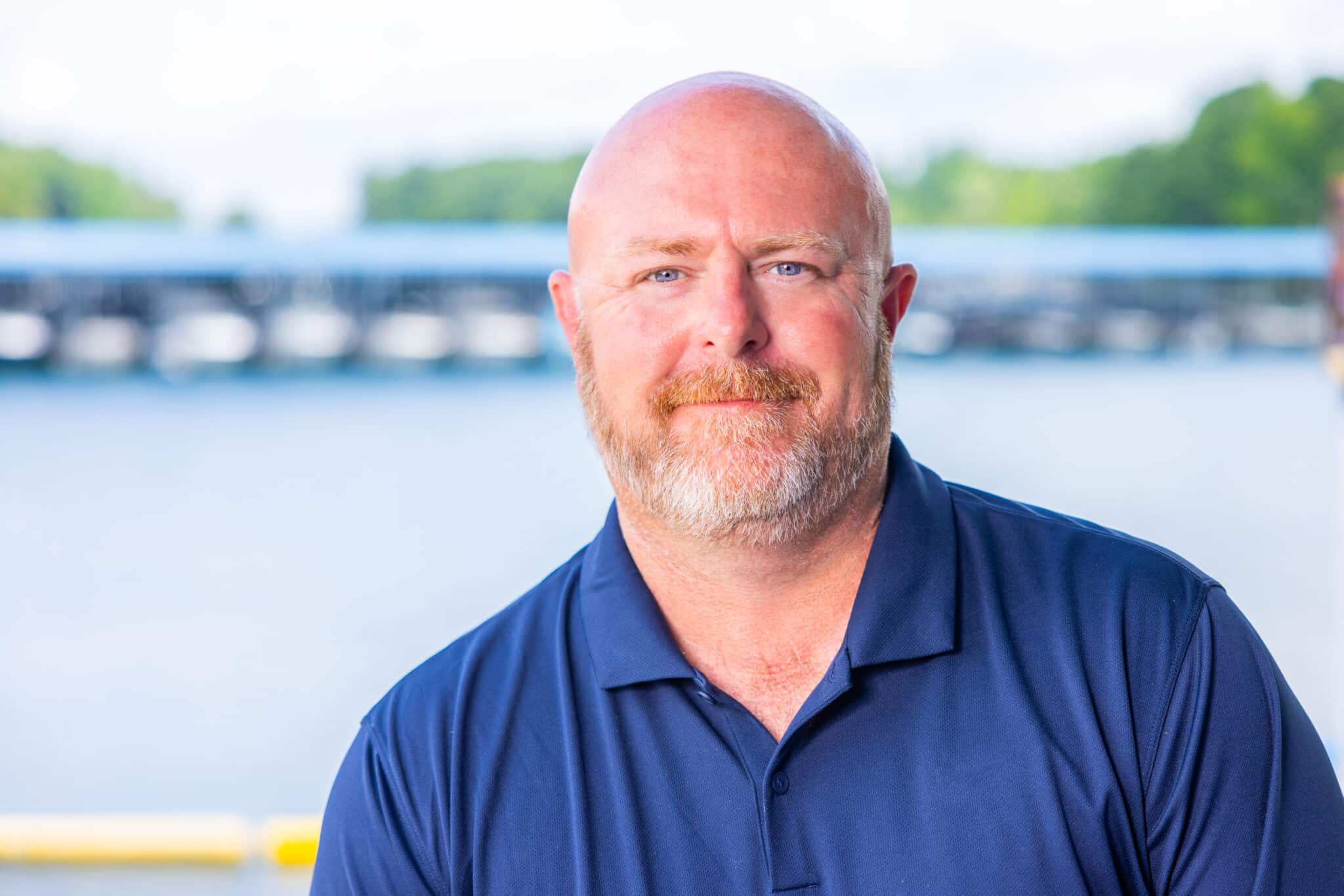 A middle-aged man with a bald head and reddish beard wears a navy blue polo shirt and stands outdoors in front of a blurred marina waterfront scene.